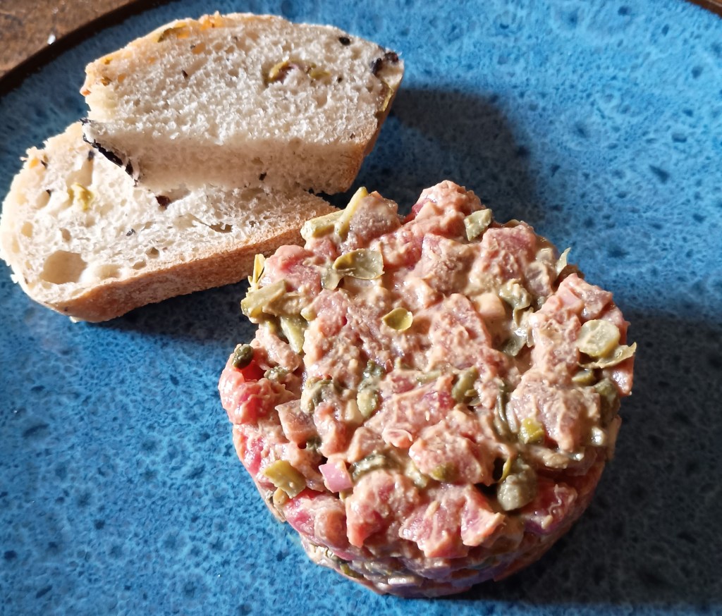 Close-up of a hand-chopped beef steak tartare, mixed with shallots, capers, cornichons, and parsley, served as a neat mound on a plate, garnished with a drizzle of olive oil, ready to be enjoyed with toasted bread.