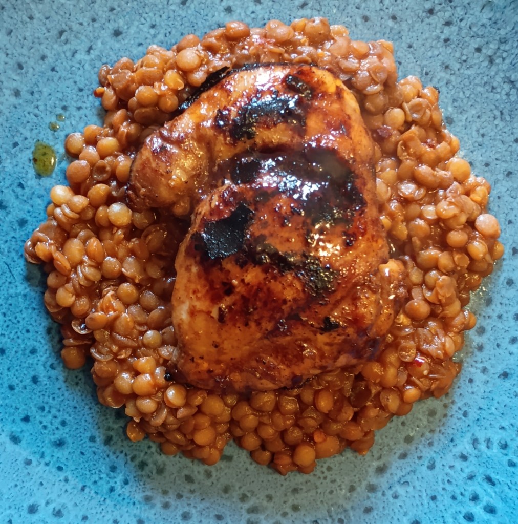 Close-up of sticky harissa and lime glazed chicken thigh served on a bed of spiced lentils, glistening with caramelised edges and rich tomato-chilli sauce, presented on a blue stoneware plate.