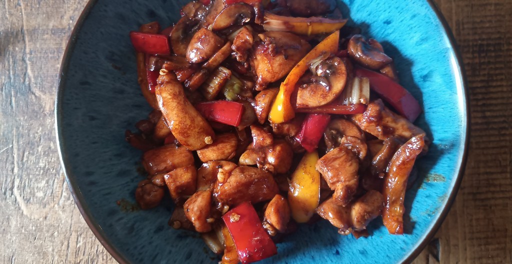A vibrant Chinese chicken stir-fry served in a deep blue bowl, featuring chunks of chicken thigh coated in glossy black bean sauce with red and yellow peppers, mushrooms, celery, and spring onions, set on a rustic wooden table.