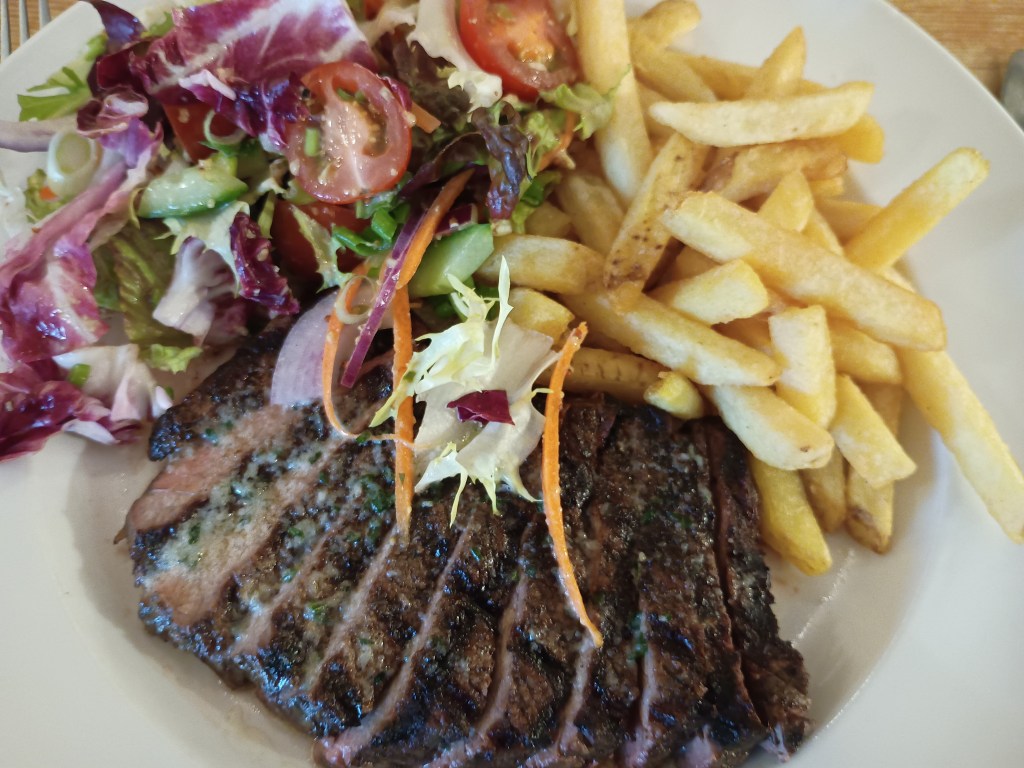 A plate of Steak Frites served at The Hare & Hounds in Fulbeck, Lincolnshire. The dish features tender, sliced steak cooked medium-rare, golden fries, and a generous helping of mixed salad with cherry tomatoes, cucumber, and leaves. Photographed close-up on a white plate, showing vibrant colours and textures.