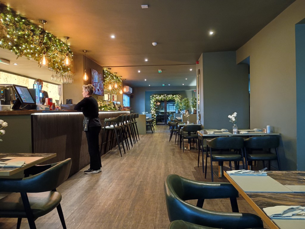 Interior of Venney’s at The Granby near North Thoresby – a stylish restaurant with modern décor, dark wood flooring, soft lighting, and greenery with fairy lights hanging above the bar area. A member of staff stands behind the bar preparing orders, while neatly set tables line the room.