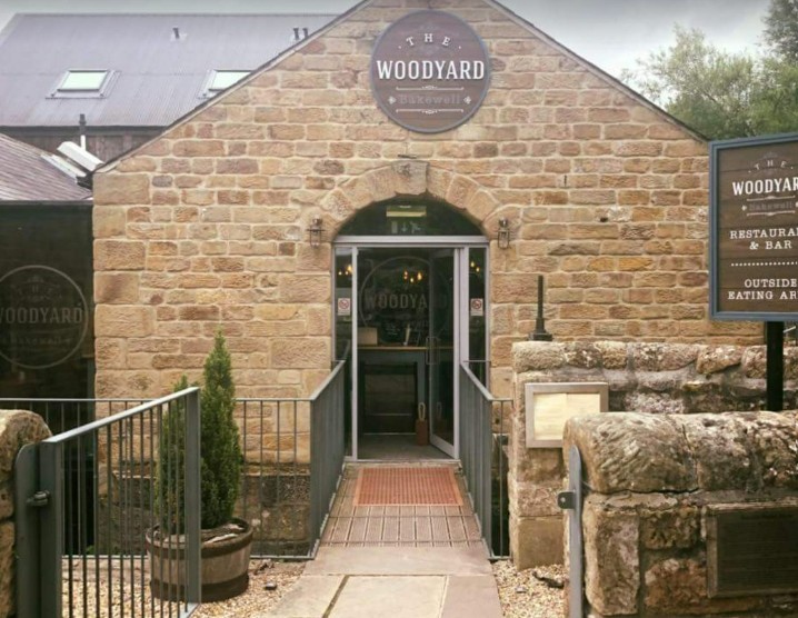 Entrance to The Woodyard restaurant in Bakewell, showing a stone building with an arched doorway, glass double doors, and signage above the entrance, with a small bridge and railings leading up to it.